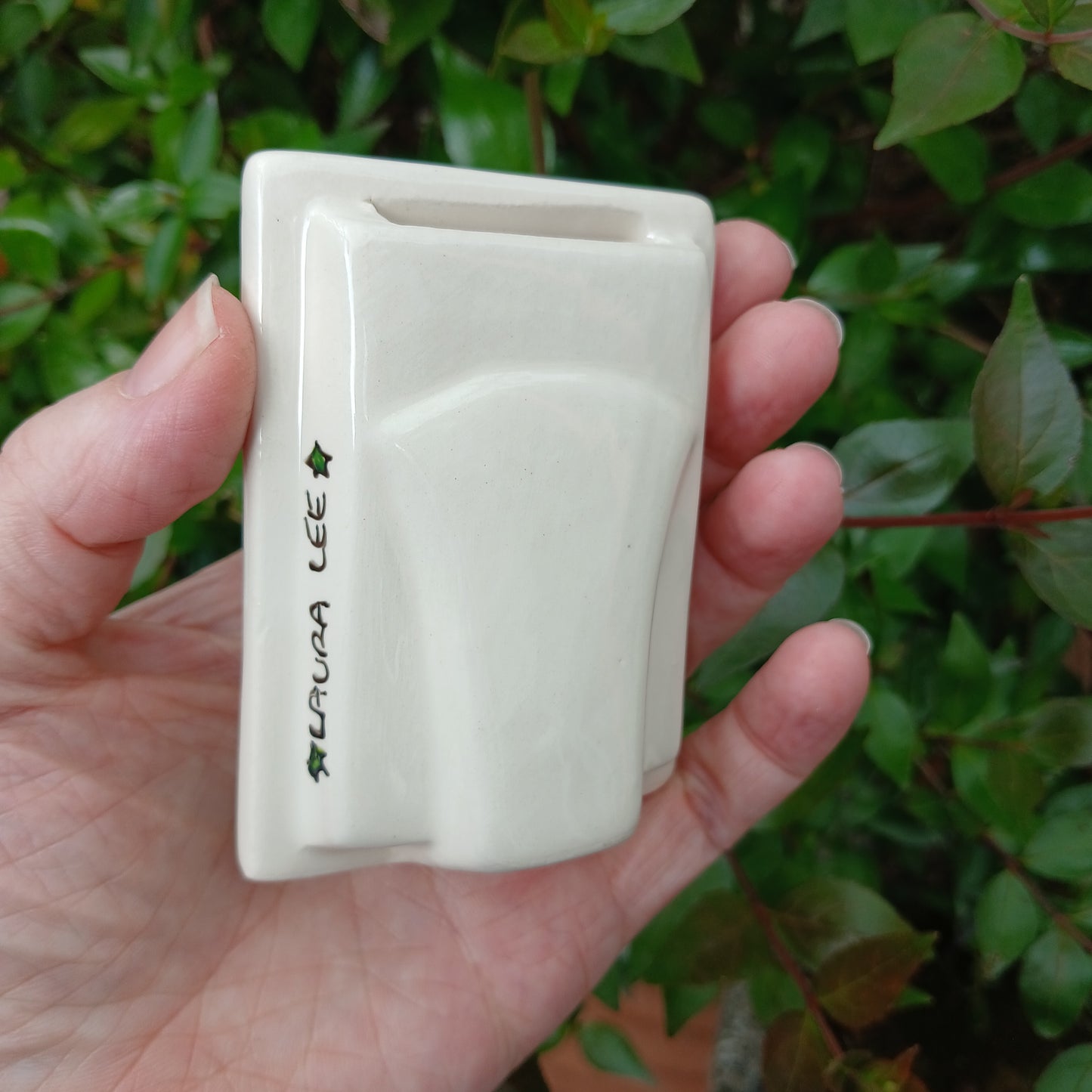Hand holding a small white ceramic photo frame signed Laura Lee with green leaves in the background