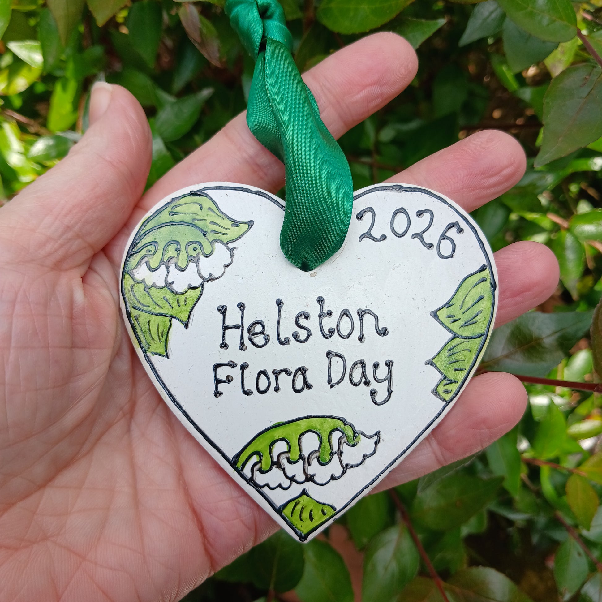 Hand holding a decorative tag with 'Helston Flora Day 2026' text against a green leafy background.