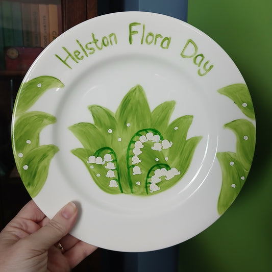 Hand-painted plate with green leaves and 'Helston Flora Day' text, held by a hand against a green background.