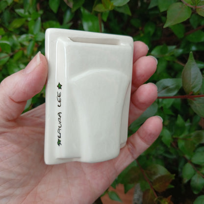 Hand holding a small white ceramic photo frame signed Laura Lee with green leaves in the background