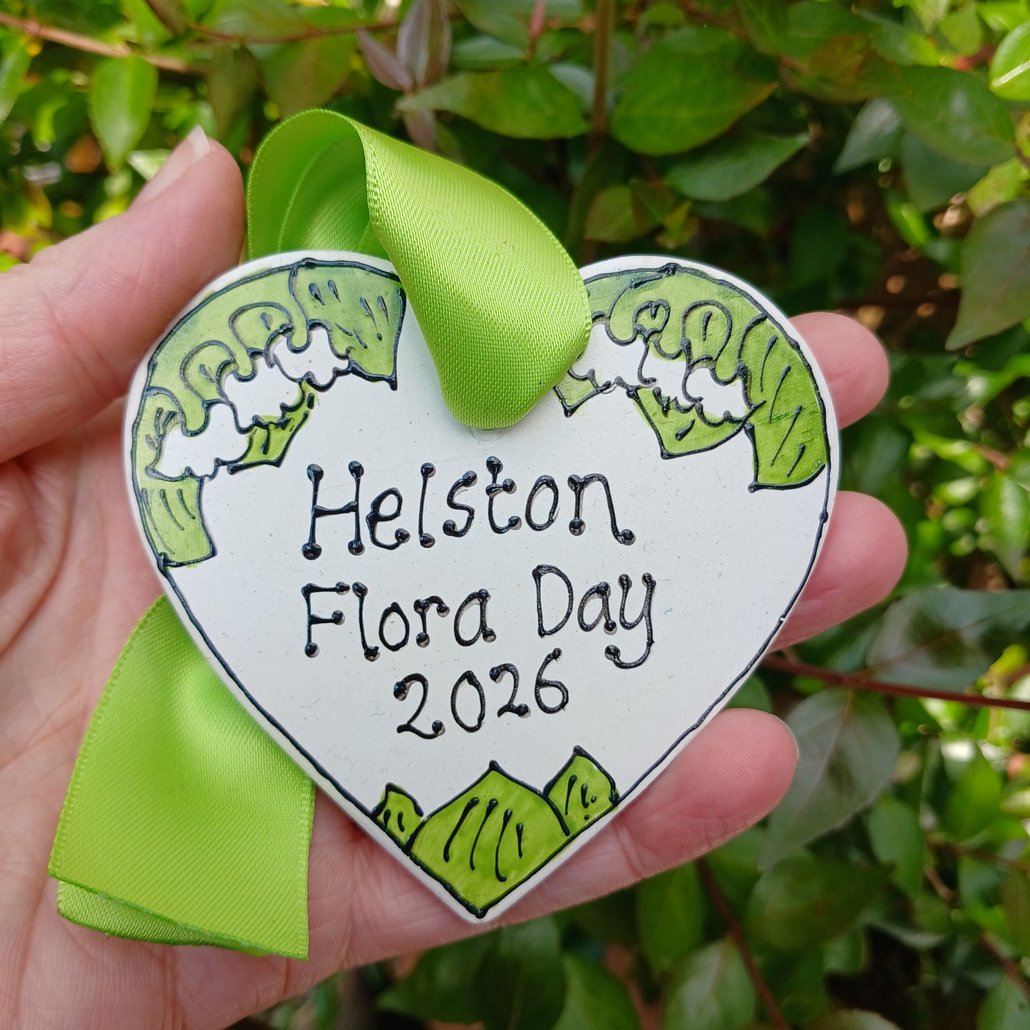 Heart-shaped badge with 'Helston Flora Day 2026' text held in a hand against a green leafy background