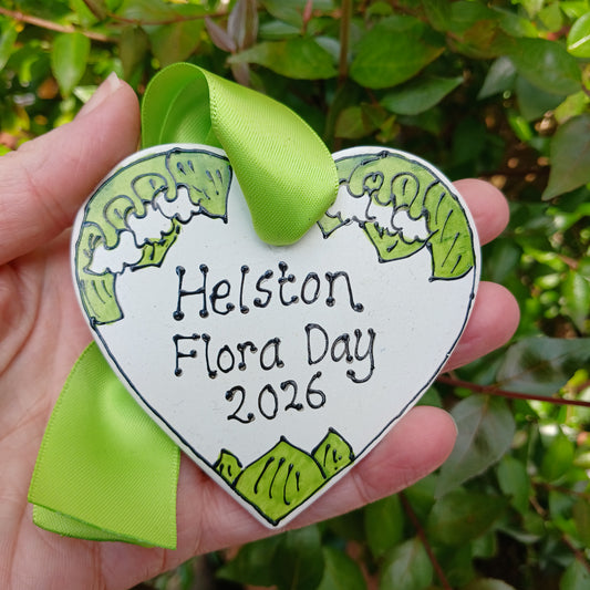Heart-shaped badge with 'Helston Flora Day 2026' text held in a hand against a green leafy background