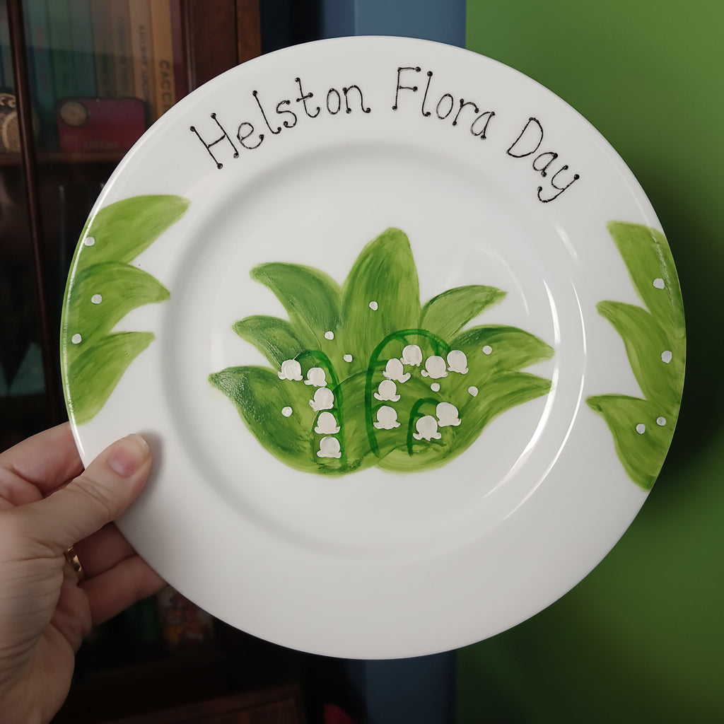 White plate with green leaf design and 'Helston Flora Day' text held by a hand against a blurred background