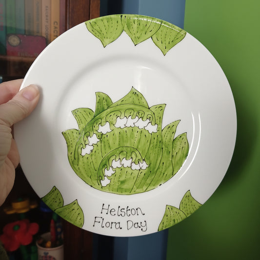 White plate with green floral design and 'Helston Flora Day' text held by a hand.