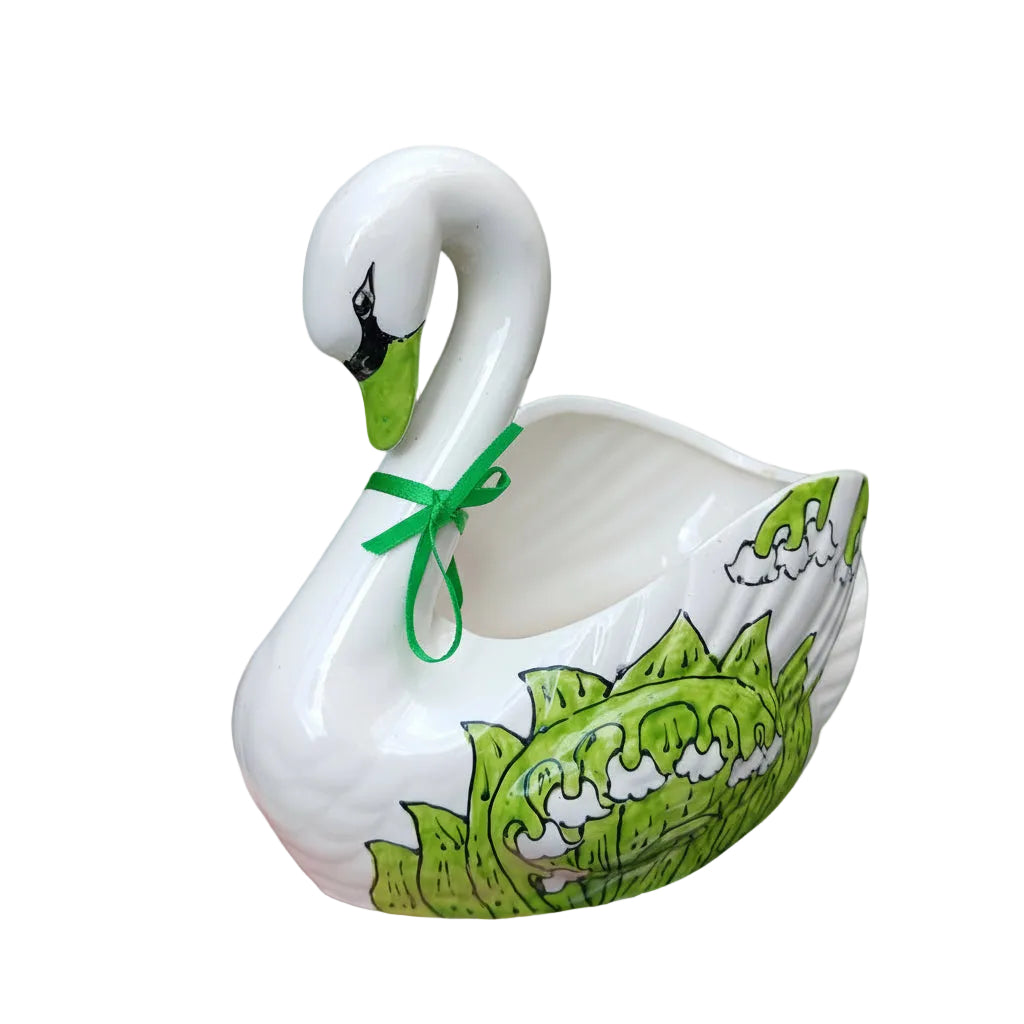 White ceramic swan with green design and bow, held against a green leafy background