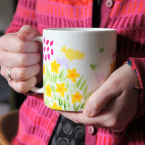 Yellow, pink, lavender, blue and orange florals with green grass tufts hand painted on a jumbo sized china mug by Cornwall based artist Laura Lee